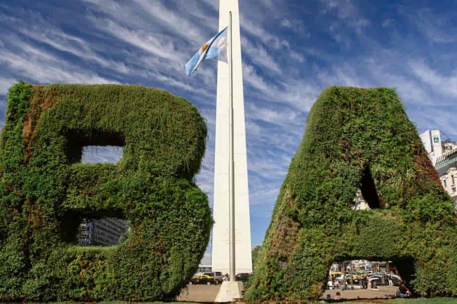 Cartel con las letras “BA” cubiertas de vegetación frente al Obelisco de Buenos Aires, con la Avenida 9 de Julio y edificios al fondo.