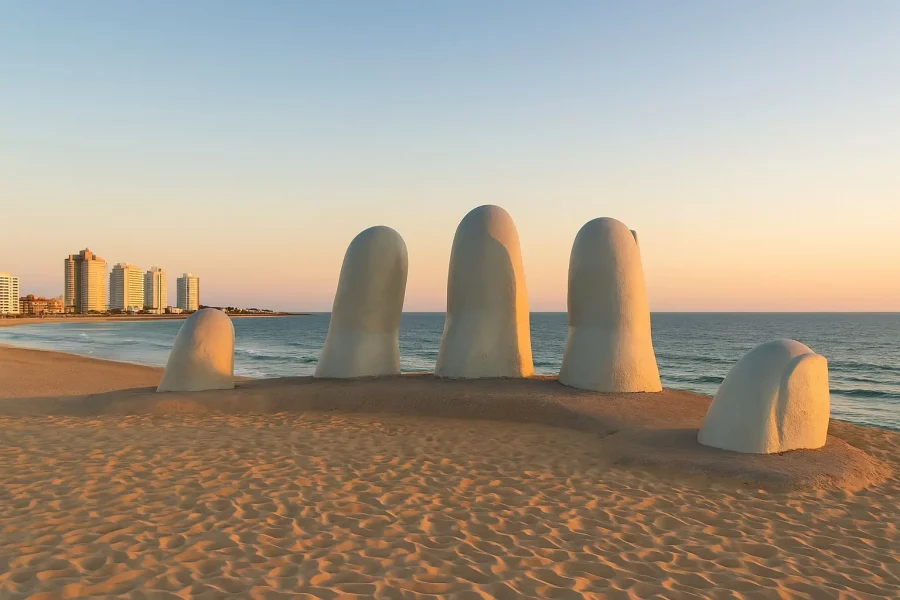 Escultura La Mano, también conocida como Los Dedos, en Playa Brava de Punta del Este al atardecer, con el mar y los edificios al fondo.